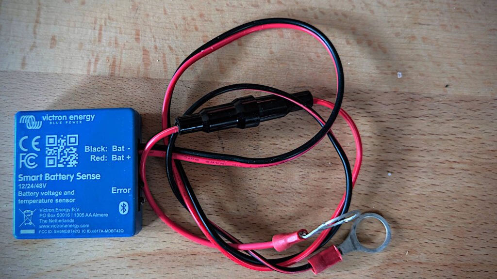 Detailaufnahme des Victron Smart Battery Sense auf einem Tisch liegend vor dem Einbau. Man sieht das kompakte blaue Gehäuse, die zwei Anschlusskabel mit Ösen für die Batteriepole und den Sicherungshalter.
